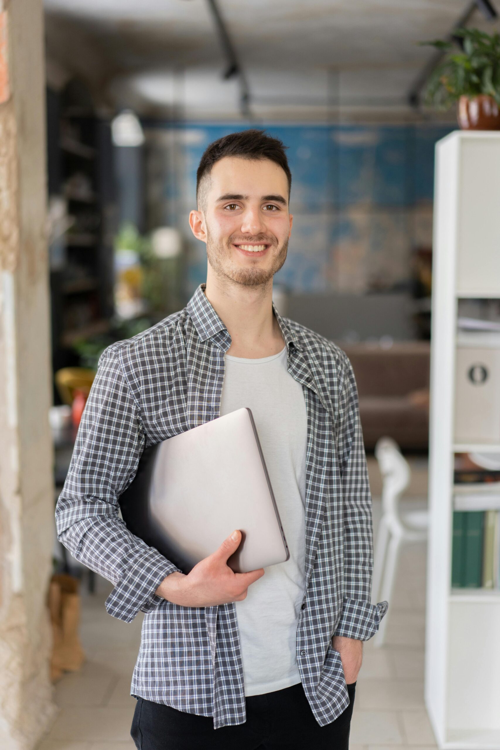Cheerful young adult man holding a laptop, standing in a contemporary office space.