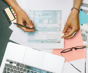 Hands writing on tax documents with laptop, glasses, and currency on desk.